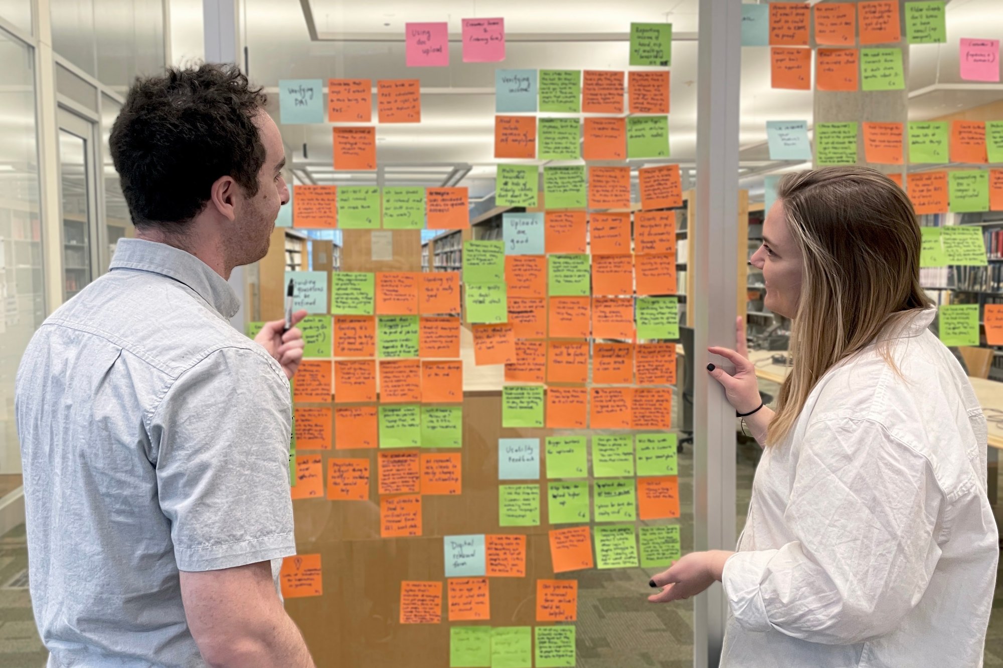 Two people having a brainstorming conversation next to a wall covered in sticky notes. 